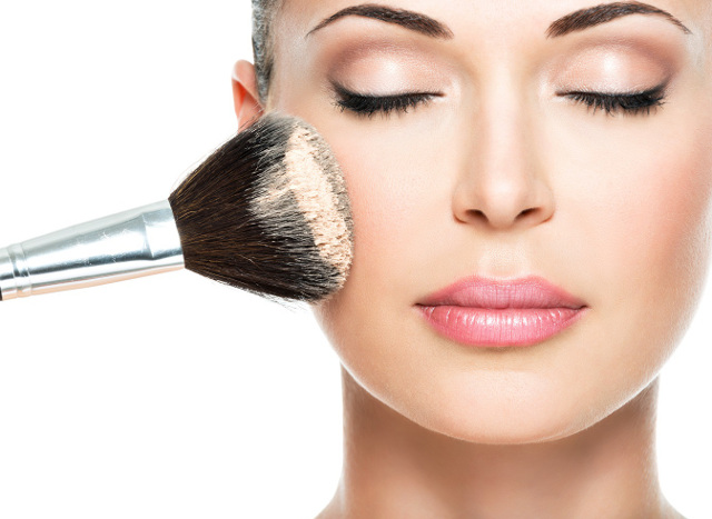 Closeup portrait of a woman applying dry cosmetic tonal foundation on the face using makeup brush.